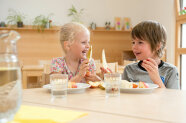 Zwei Kinder sitzen hinter Tellern und Gläsern an Tisch und halten Birnenspalte in der Hand